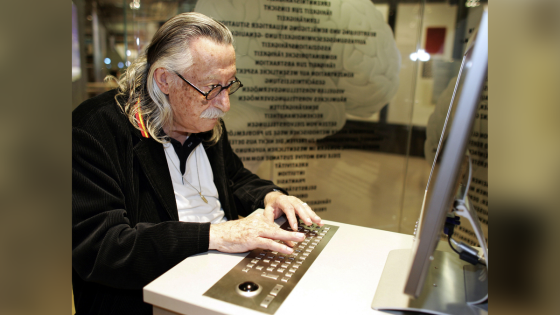 An older white man with long salt-and-pepper hair and glasses sits at a desk with a keyboard and computer monitor