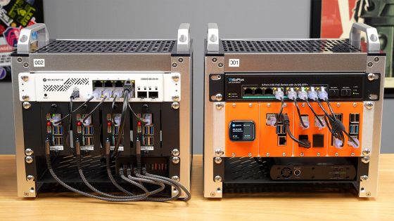 Two mini-racks in cube-style silver containers, one with an orange back and the other silver, on a wooden desk, ethernet cables neatly fanning out from each.