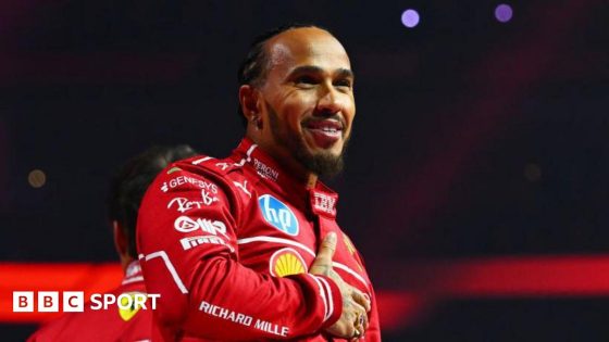 Lewis Hamilton holds his right hand across his chest while smiling at the F1 2025 season launch event. He is wearing the red Ferrari uniform 