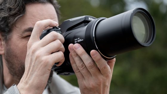 A person is holding a Nikon camera with a large lens, capturing a photo. The individual appears focused, with the lens pointed forward. The background is softly blurred with greenery.