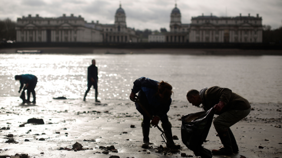 Four backlit people bend over and pick up items from the beach of the Thames at low tide, with buildings in the background