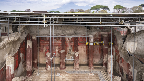 View from above of a newly excavated room at Pompeii; there are columns close to the interior walls, which are painted red with images of people and mythical beings. Vesuvius rises in the background.