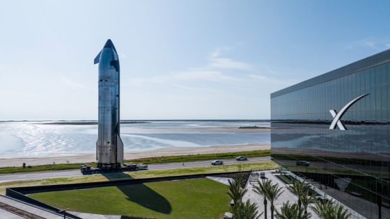 a silver winged starship ship is moved to launch pad with SpaceX building in foreground