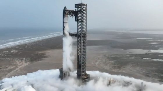 A giant silver Starship rocket is stacked atop its giant booster atop a seaside launch pad for SpaceX