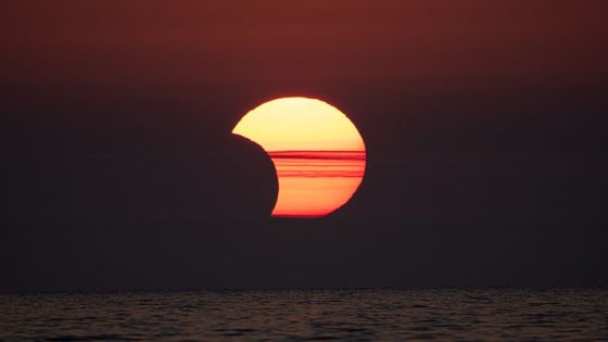 partial solar eclipse showing the moon appearing to take a 'bite' out of the sun behind some hazy clouds above the ocean. 