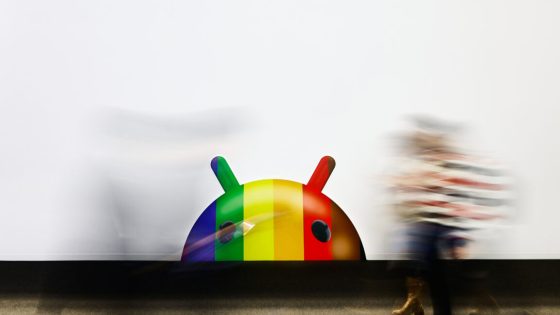 Android robot mascot, its head in rainbow stripes, peeking above a white background horizon, with people blurred in motion moving past.
