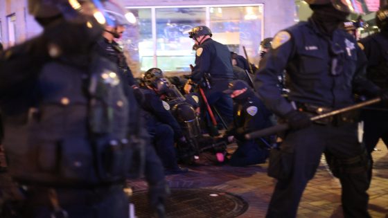 A group of police officers and ICE agents in uniform and protective gear detain an individual on a city sidewalk at night.
