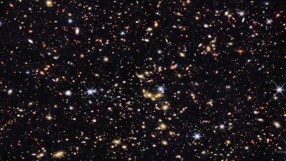 An area of deep space with thousands of galaxies in various shapes and sizes on a black background. Most are circles or ovals, with a few spirals. More distant galaxies are redder in color and smaller, down to being mere dots, while closer galaxies are a bit larger and white or blueish. A few gold-colored galaxies are bunched closely together in the center. Bright stars surrounded by spikes lie in our galaxy. Credit: NASA/ESA