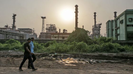 Pedestrains walk past an oil refinery operated by Bharat Petroleum Corp. Ltd. with the sun shining thorugh a very overcast sky in the background, in Mumbai, India.