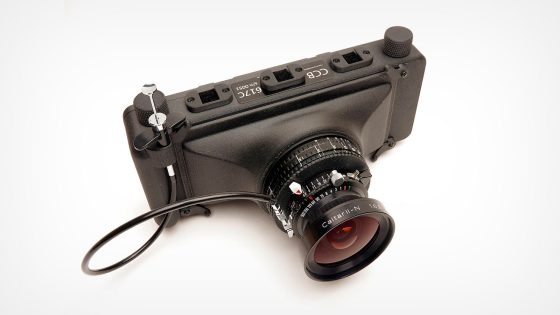A black, compact digital camera with several labeled ports on top and a wide-angle lens attached; a cable release is connected to the camera. The camera is photographed on a white background.