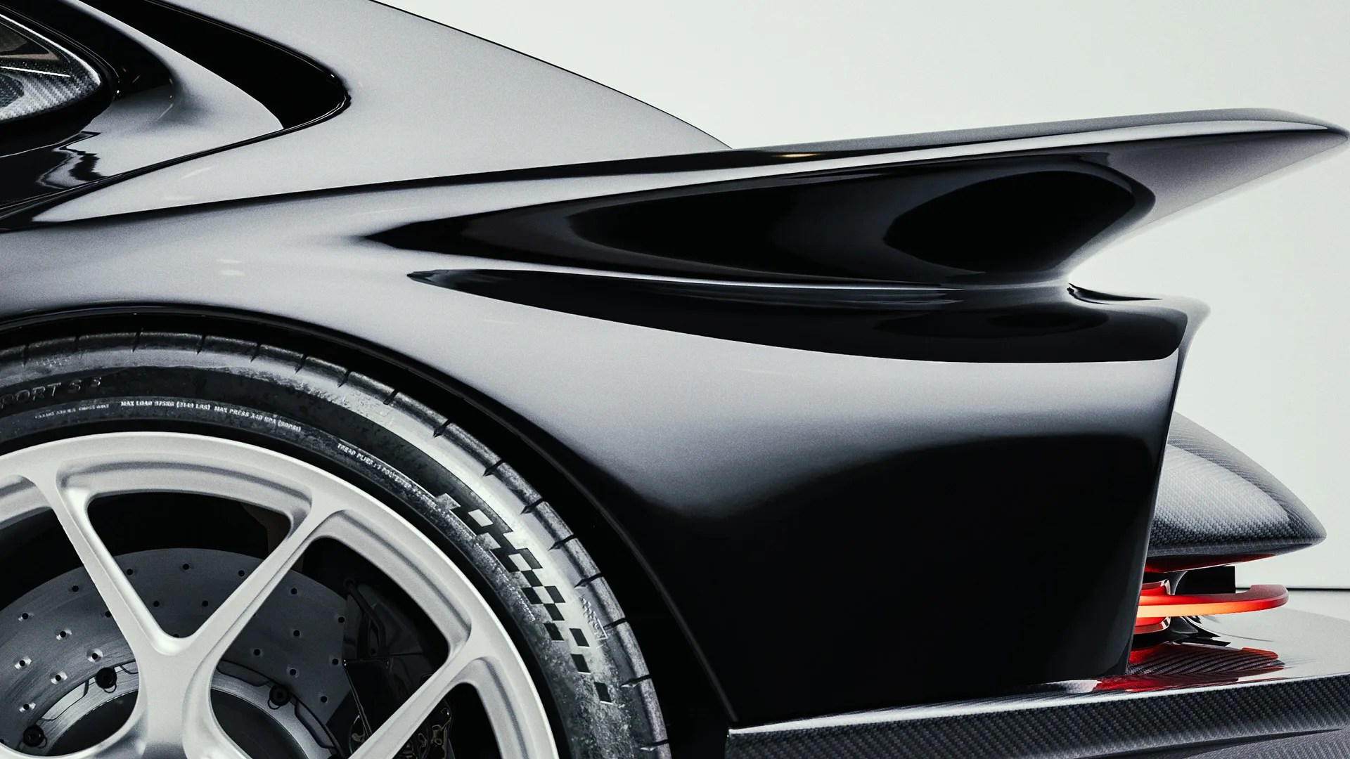 Close-up of the rear section of a sleek black sports car, highlighting the aerodynamic rear wing, part of the rear wheel with a silver rim, and a carbon fiber diffuser. The car